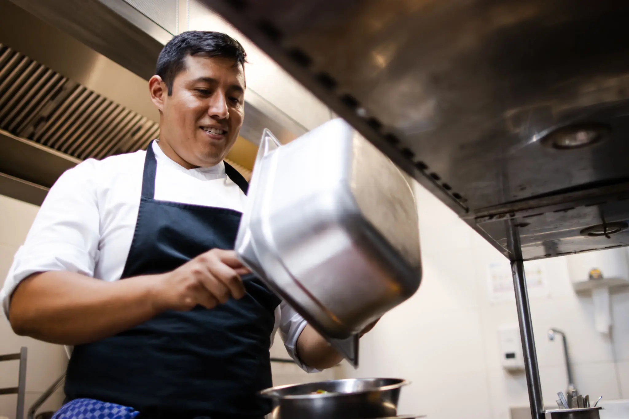 Chef pouring sauce from a saucepan in a kitchen.