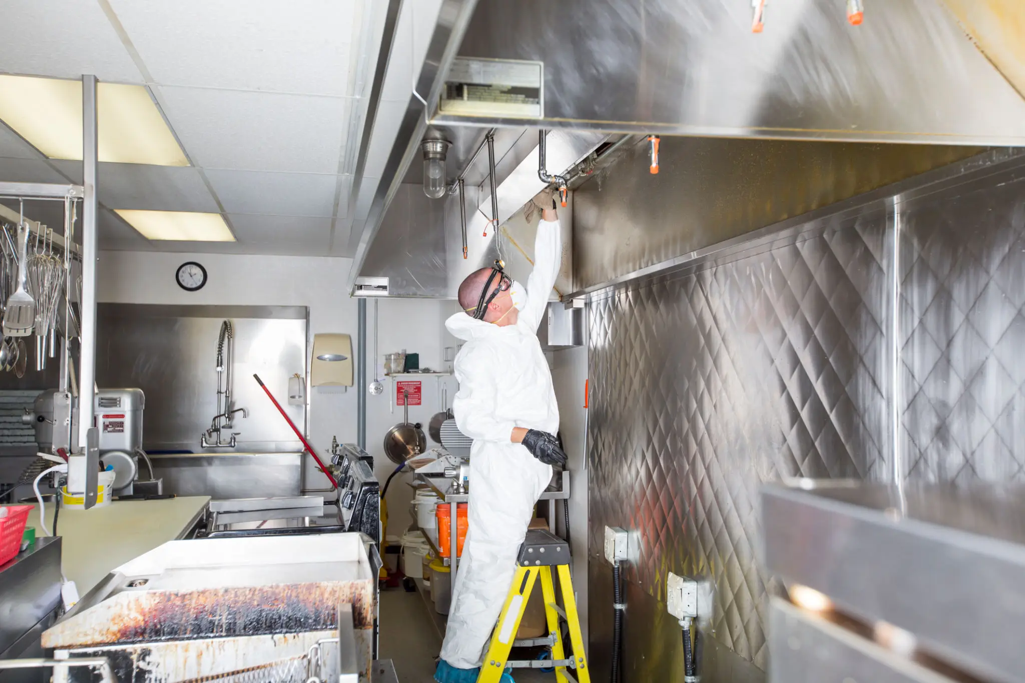 Worker on a ladder fixing kitchen equipment in a commercial kitchen.